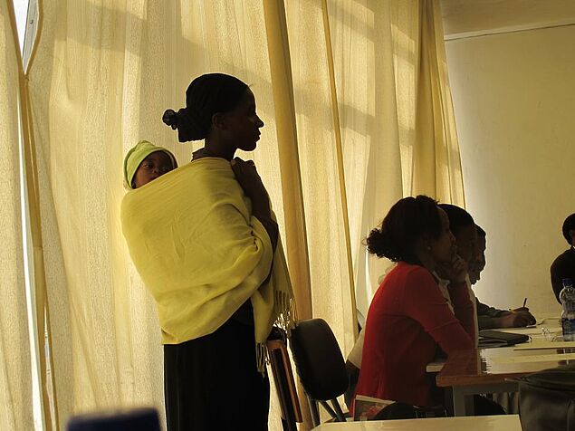 Woman with baby standing and other parents sitting at desks listening.